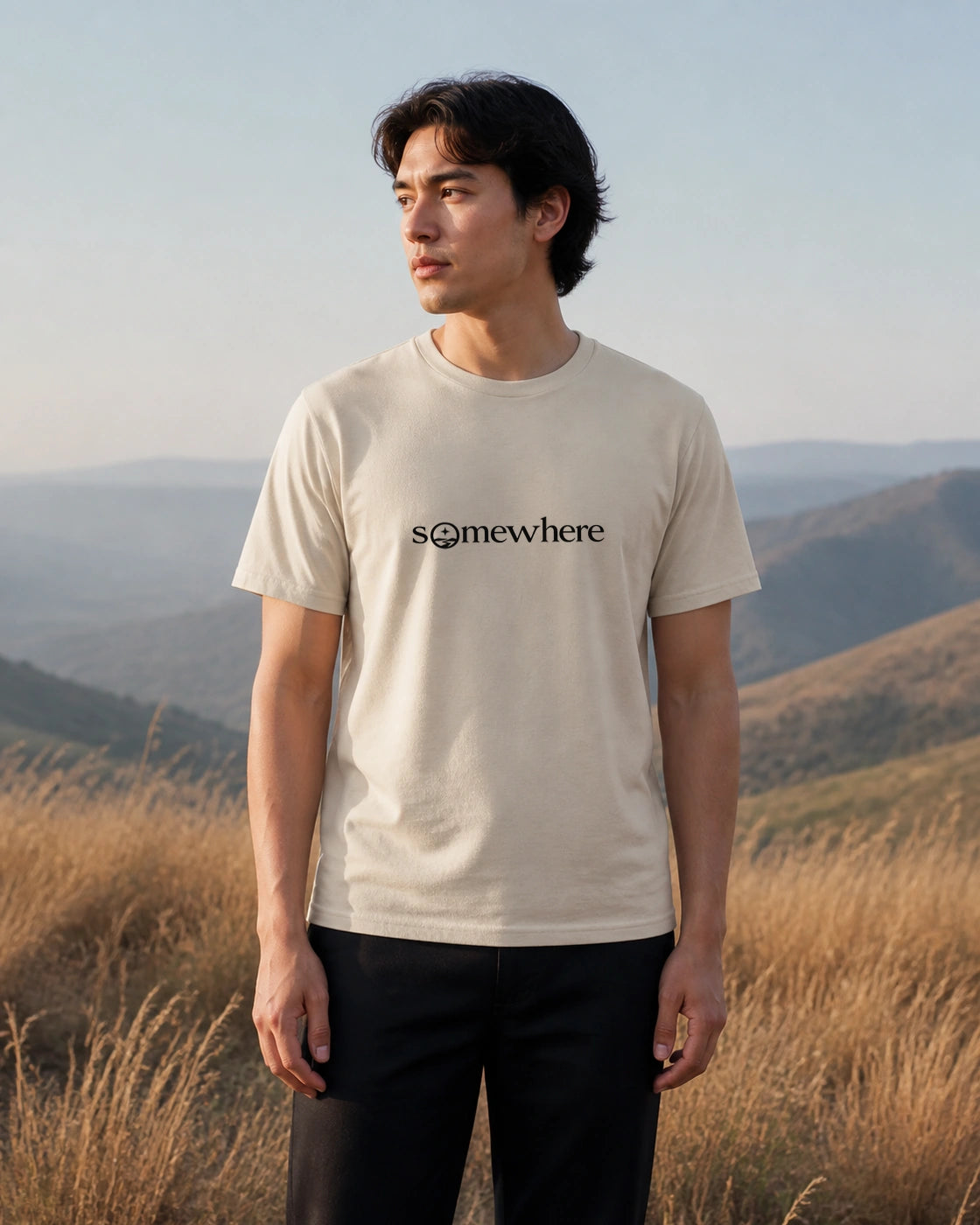 Model wearing the Somewhere wordmark tee in Sand outdoors in a mountain landscape